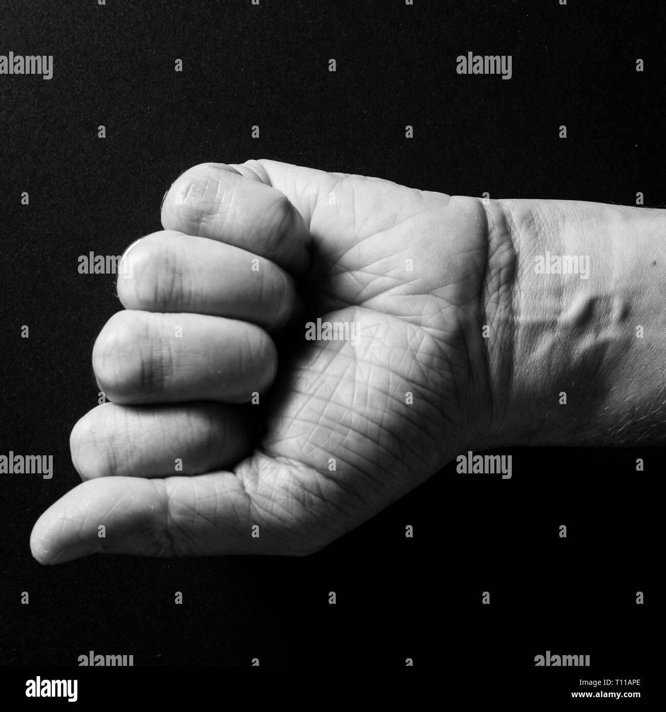 Black & white image of man`s clenched fist and wrist, isolated against a black background with dramatic sidelight Stock Photo